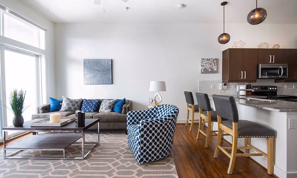 A modern living room and kitchen area with a gray sofa adorned with blue and patterned pillows, a blue patterned armchair, a wooden coffee table with decorative items, and a kitchen counter with four bar stools. The room has wooden flooring, white walls, and large windows allowing natural light to enter.