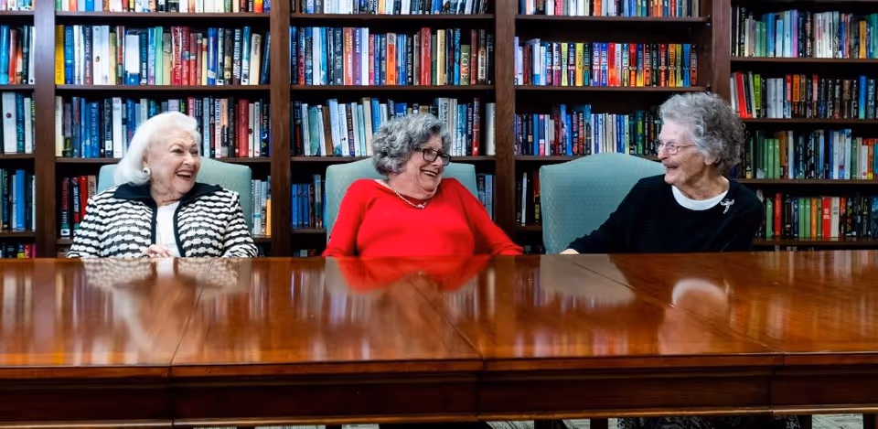 Three elderly women sitting at a wooden table in front of a large bookshelf filled with books, smiling and engaging in conversation.