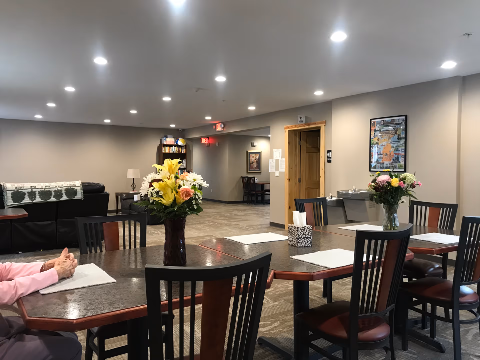 A well-lit common area in South Torch Assisted Living featuring a large table with chairs around it, two vases with colorful flowers on the table, a water fountain on the wall, a bookshelf, a couch with a blanket, and a person sitting at the table with hands folded.