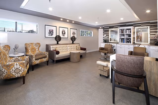 A spacious and well-lit salon area in a senior living facility featuring two patterned armchairs, a tufted white sofa with two hair drying stations behind it, and salon styling chairs in front of mirrors with shelves and cabinets. The walls are decorated with framed artwork and the floor is covered with a neutral-toned carpet.