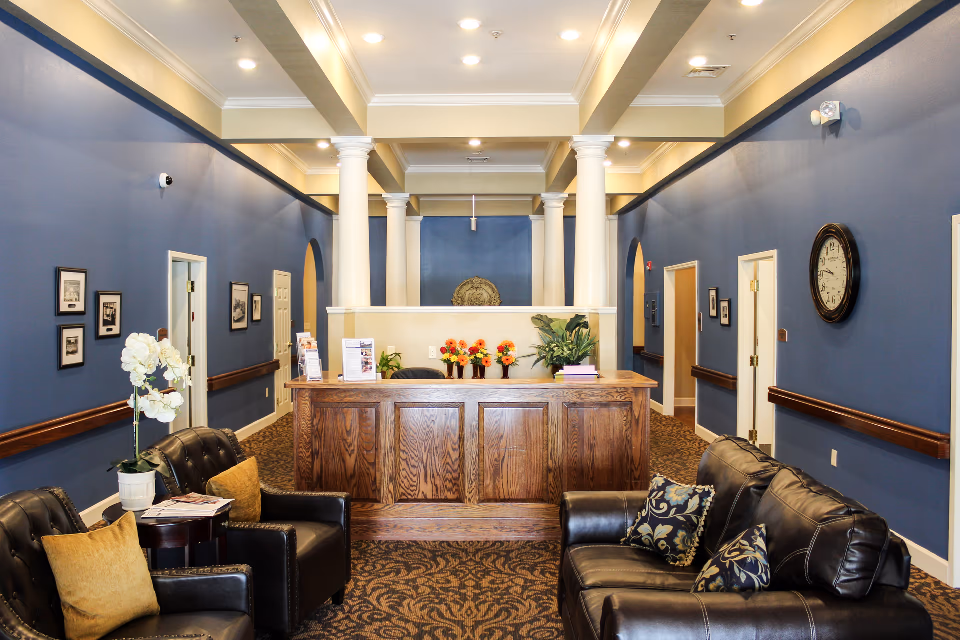 Reception area with a wooden front desk adorned with flowers and plants, surrounded by blue walls with white columns and several doors. There are black leather chairs and a sofa with decorative pillows in the foreground, a wall clock, framed pictures, and a patterned carpet.