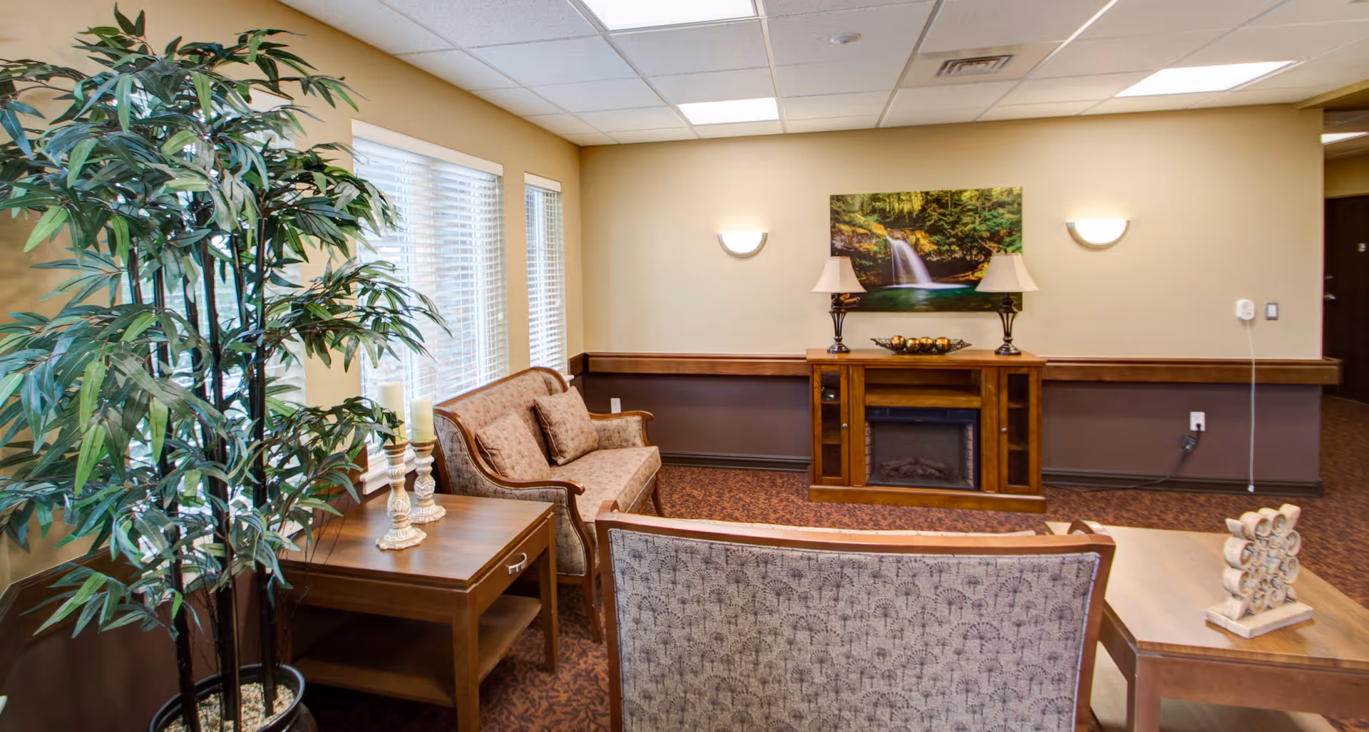 A cozy living room area with two upholstered sofas facing a wooden electric fireplace. There is a large potted plant next to a side table with decorative candles. Above the fireplace is a framed picture of a waterfall in a forest. The room has beige walls, carpeted floor, and ceiling lights.