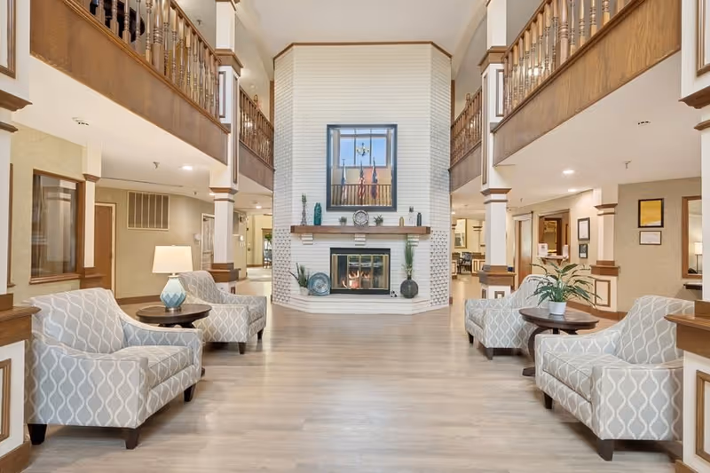 A spacious and well-lit living room area with a white brick fireplace in the center. The room features four patterned armchairs arranged around two small round wooden tables, one with a lamp and the other with a potted plant. The space has light wood flooring, beige walls, and wooden railings on the upper level balcony overlooking the room. Decorative items are placed on the fireplace mantel, and there is a large window above the fireplace.