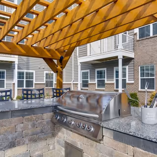 Outdoor grilling area with a stainless steel built-in gas grill under a wooden pergola. The area features granite countertops and stone walls, with a residential building visible in the background.