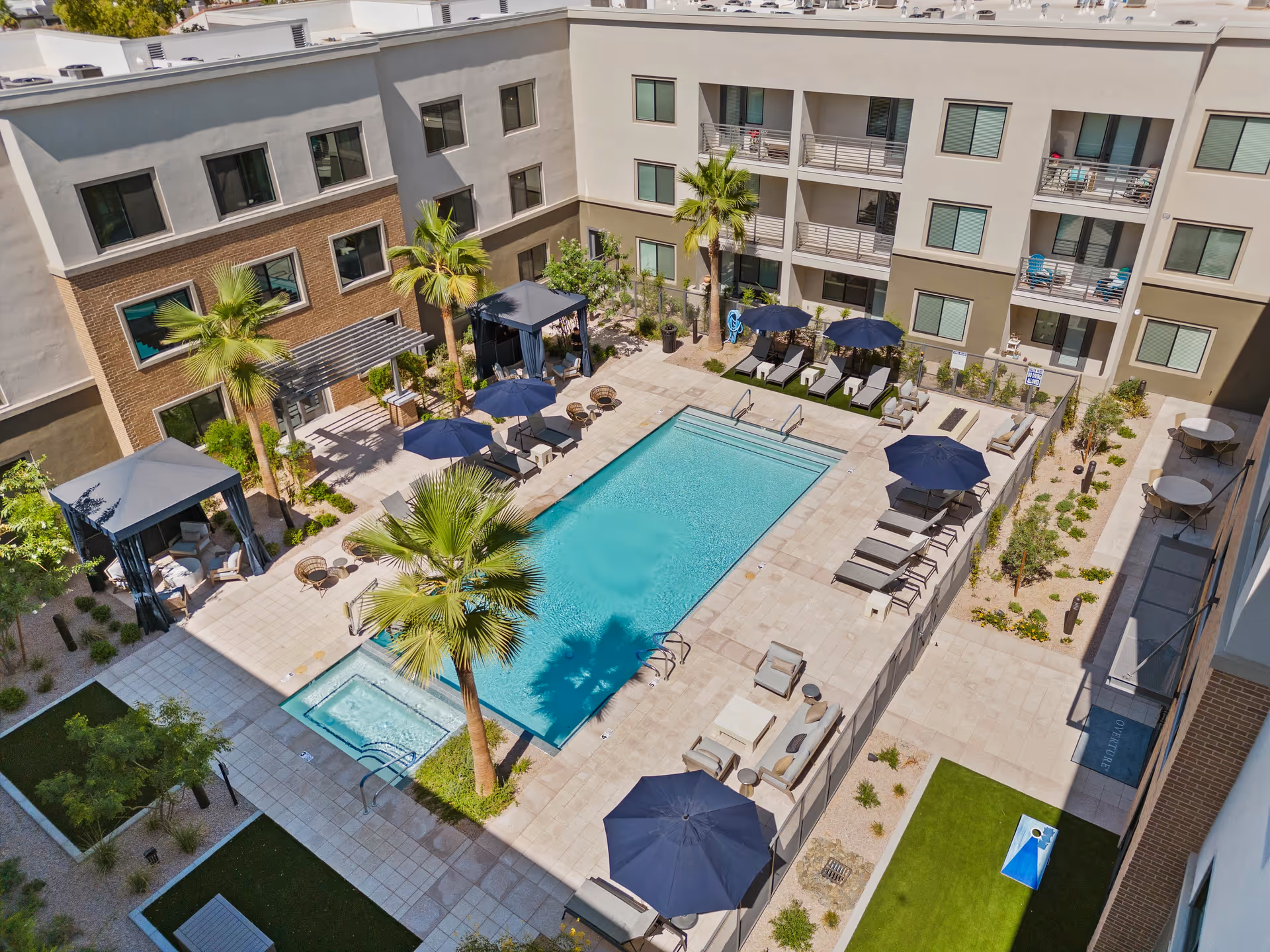 Aerial view of a sunlit courtyard with a rectangular swimming pool, hot tub, lounge chairs, umbrellas and cabanas surrounded by a multi-story building.