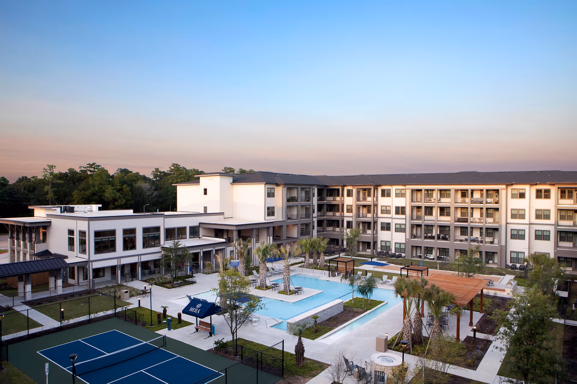 Outdoor view of Mera Vintage Park senior living facility featuring a large swimming pool surrounded by palm trees, lounge chairs, and shaded seating areas. There is a tennis court in the foreground and a multi-story residential building in the background under a clear sky at dusk.