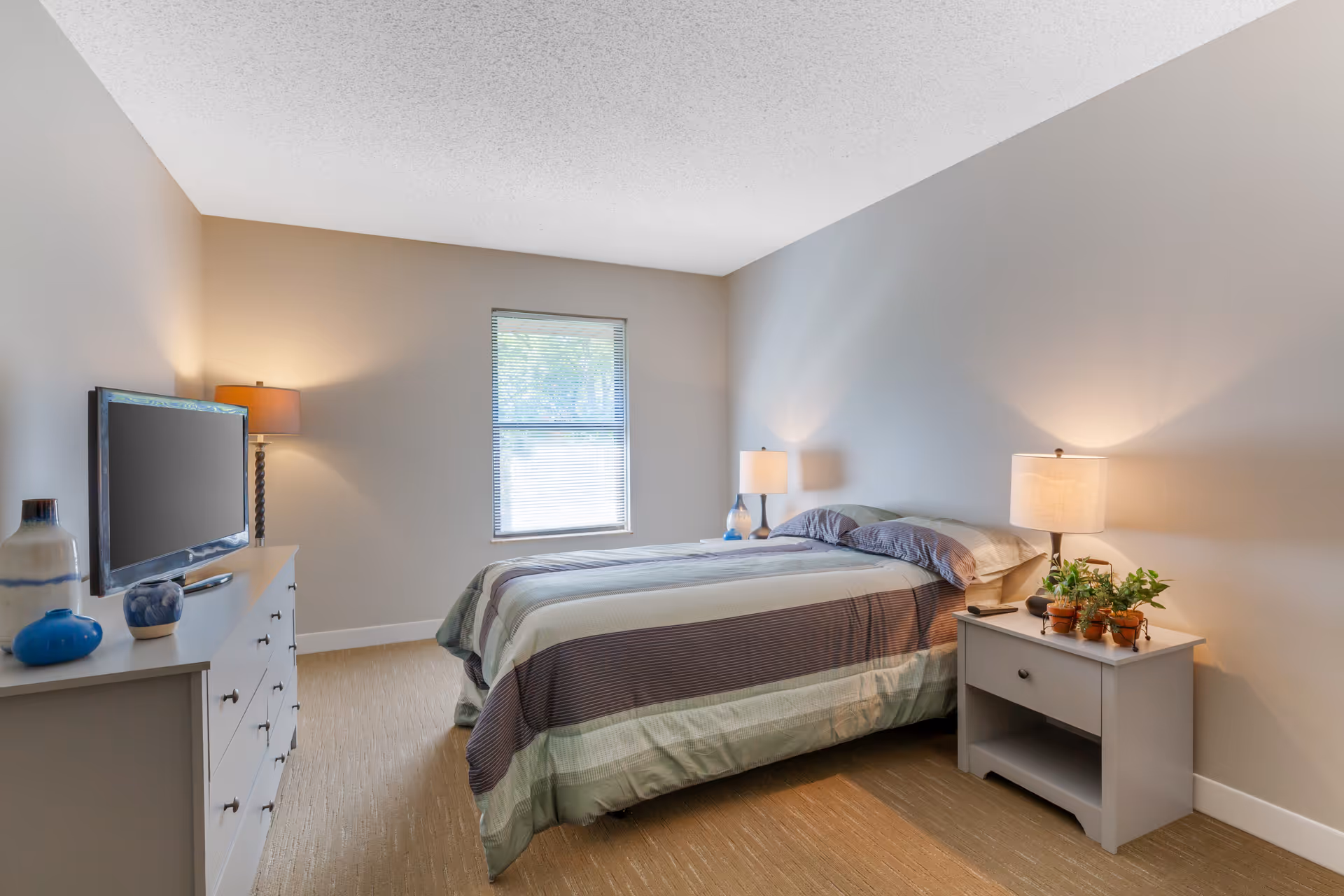 A neatly arranged bedroom with a bed covered in a striped comforter, two bedside tables each with a lamp, and a dresser with decorative vases and a flat-screen TV. There is a window with blinds letting in natural light.