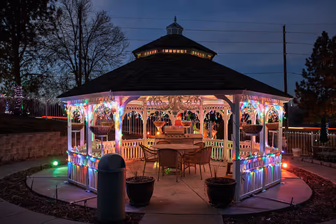 A white wooden gazebo decorated with colorful string lights at dusk, surrounded by a paved walkway and some trees in the background. Inside the gazebo, there are chairs and a table with a piano, and hanging baskets are visible on the gazebo's posts.