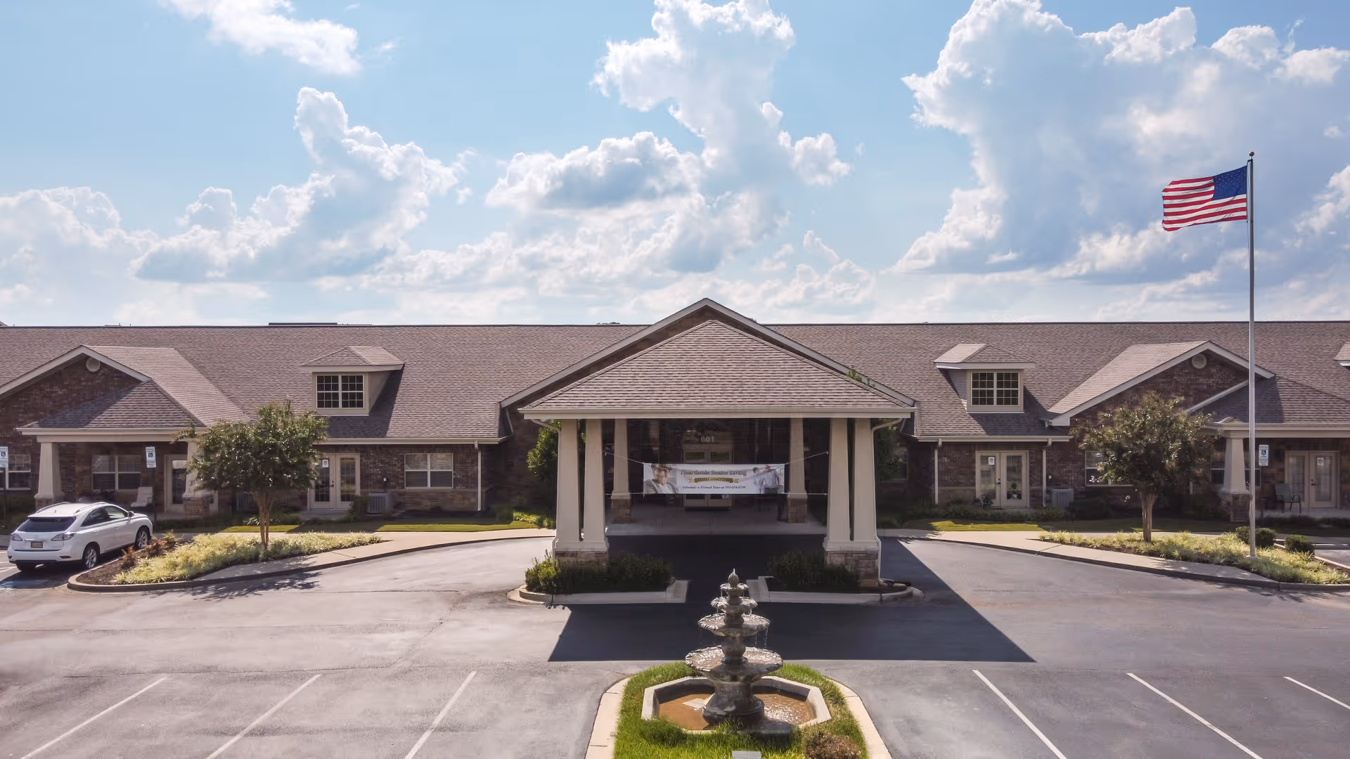 Front exterior view of Hearthside Senior Living facility with a covered entrance, a three-tiered fountain in the driveway, an American flag on a flagpole, and a partly cloudy sky.