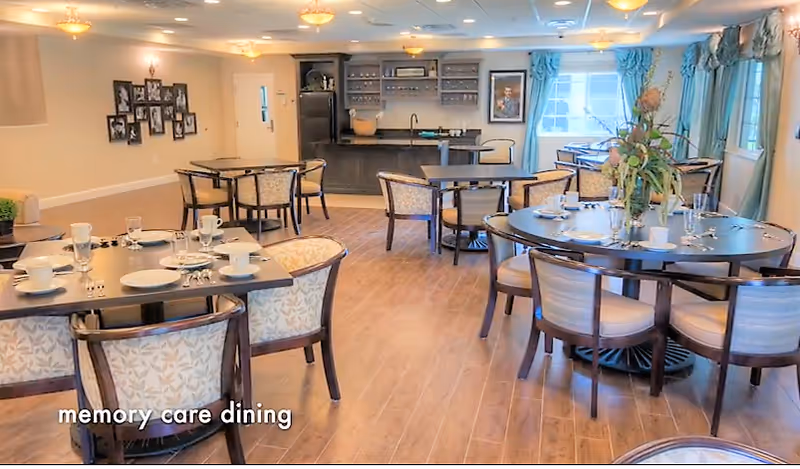 Bright memory-care dining room with multiple round and rectangular tables set for meals and a serving area and windows in the background.