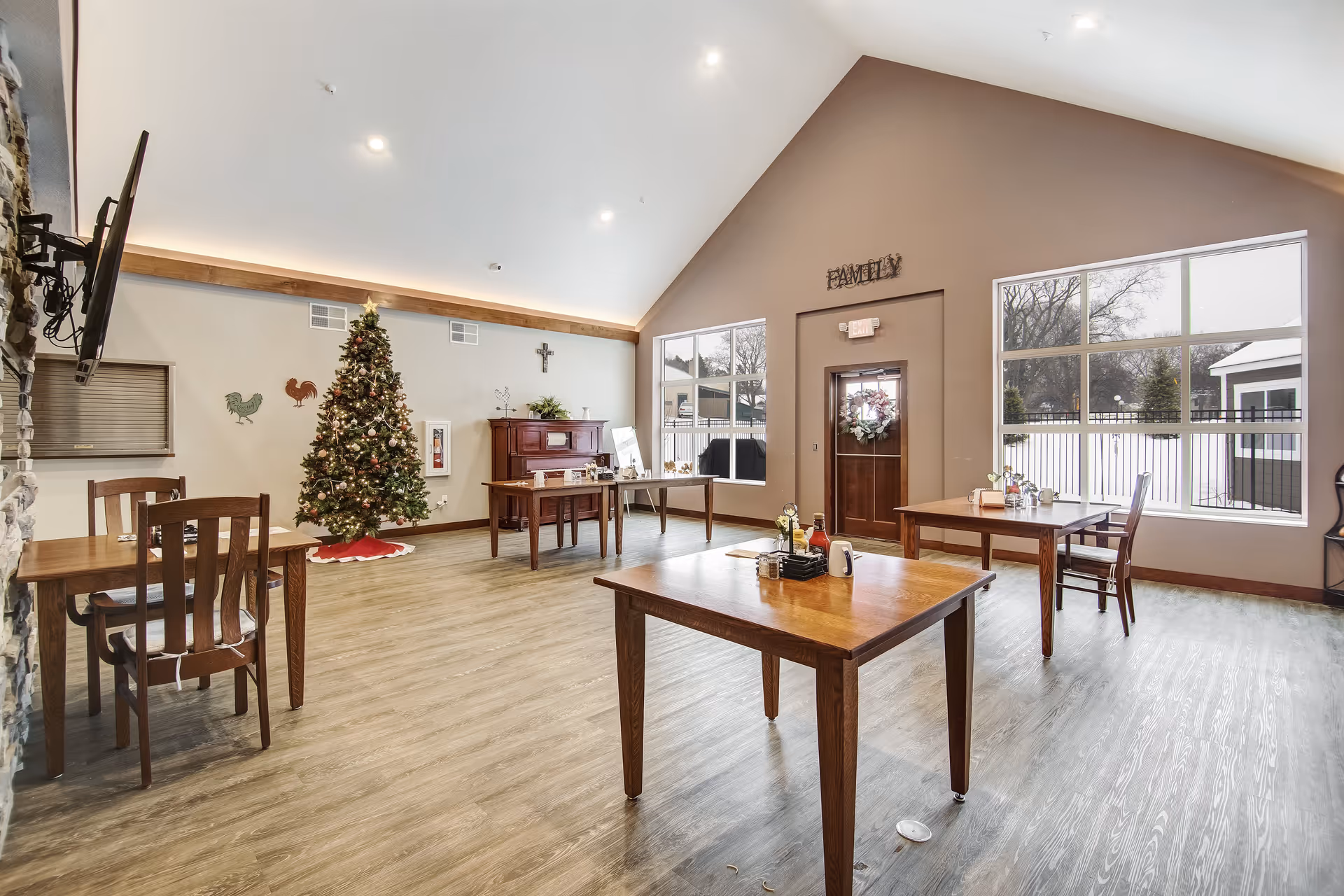 A spacious dining room with wooden tables and chairs, decorated with a Christmas tree and a wreath on the door. Large windows let in natural light, and a piano and wall decorations are visible in the background.