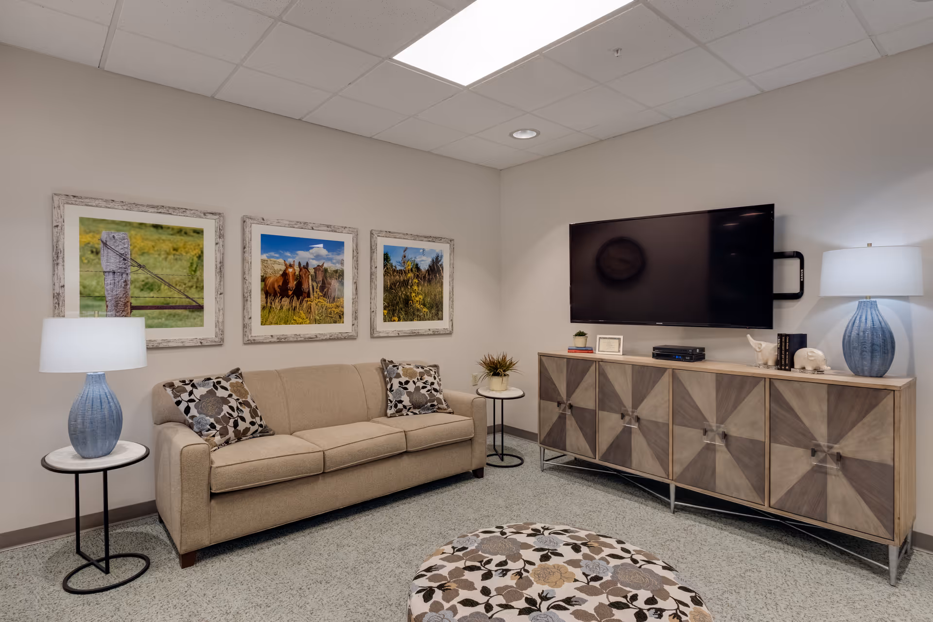 A cozy living room area with a beige sofa adorned with floral patterned pillows, two blue table lamps on round side tables, a large flat-screen TV mounted on the wall above a wooden cabinet with geometric patterns, and three framed nature photographs hanging on the wall behind the sofa.