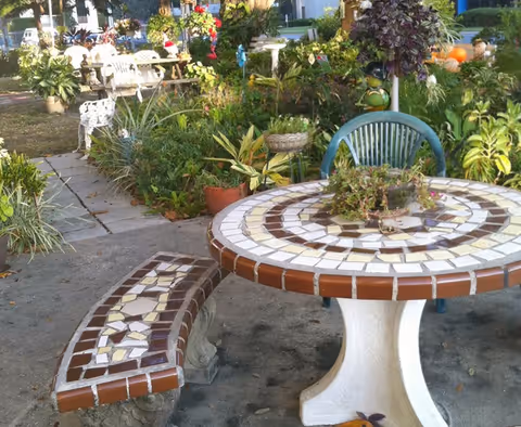 Mosaic-tiled round outdoor table with a matching bench and a plastic chair surrounded by plants and garden decorations.