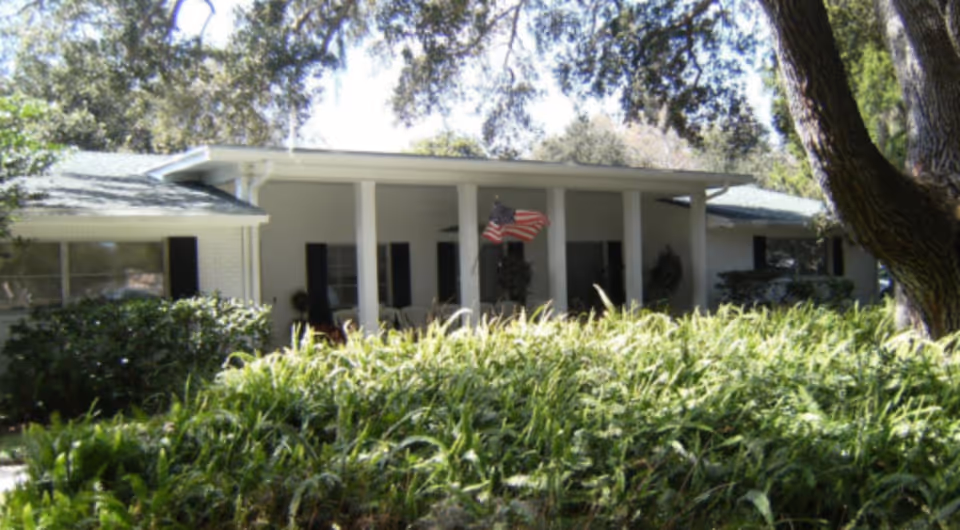 Exterior view of a single-story building with white walls and a green roof, partially obscured by tall green plants and trees. The building has a covered porch with white columns and an American flag hanging in the center.