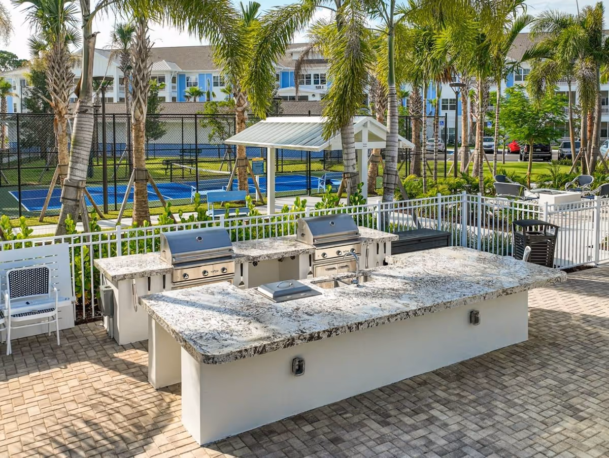 Outdoor communal grilling area with granite countertops and stainless steel grills, palm trees, seating, and a fenced sports court in the background.