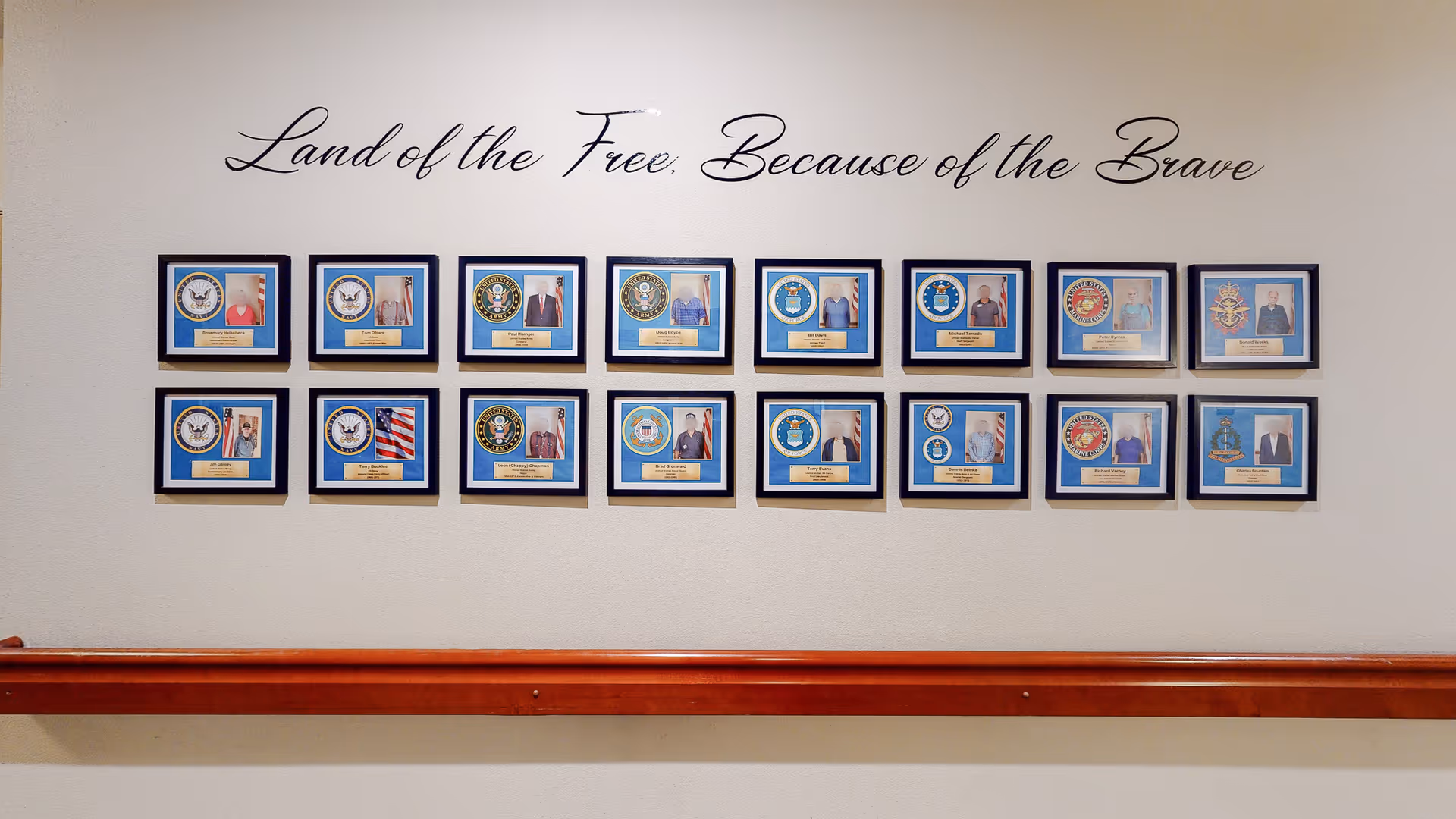 A wall display featuring framed photos and military insignias honoring veterans, with the phrase 'Land of the Free. Because of the Brave' written above. The display is mounted above a wooden handrail.