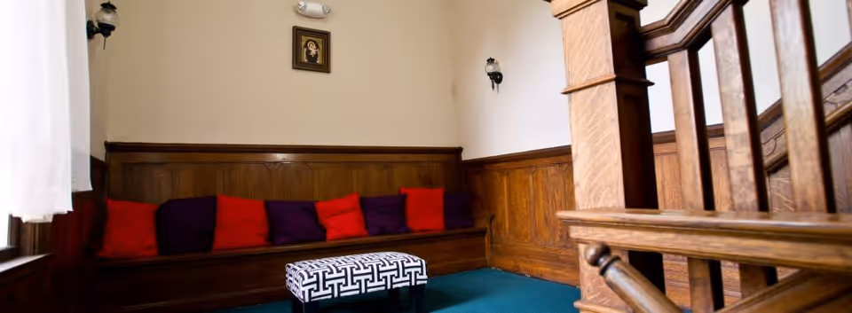 A cozy interior corner with wooden paneling and a built-in bench lined with red and purple cushions. A small patterned ottoman is placed in front of the bench, and a wooden staircase railing is visible on the right side. The walls are light-colored with two wall-mounted lamps and a small framed picture above the bench.