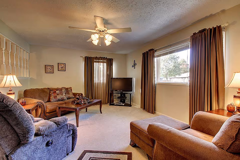 A cozy living room with beige walls and carpet, featuring a brown sofa with patterned cushions, a matching armchair with an ottoman, a wooden coffee table with a decorative centerpiece, a flat-screen TV on a black stand, two windows with brown curtains, and two table lamps on wooden side tables providing warm lighting.