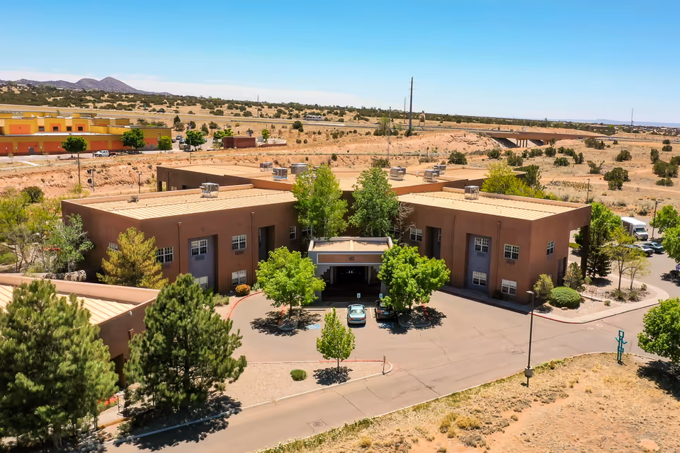 Aerial view of a brown, two-story building with a flat roof surrounded by trees and a parking area with a few cars. The building is located in a dry, desert-like landscape with sparse vegetation and distant hills under a clear blue sky.