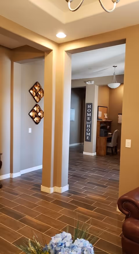 Interior view of a living space with beige walls and brown tiled flooring. There is a decorative wall art piece with four diamond-shaped panels on the left wall. In the background, a hallway leads to a small office area with a desk, chair, and a sign on the wall that reads 'HOME Sweet HOME'. A portion of a brown leather armchair and a floral arrangement with blue flowers are visible in the foreground.