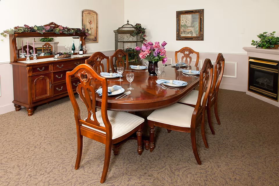A formal dining room with a polished wooden dining table set for six, featuring white plates, folded napkins, and wine glasses. The room includes matching wooden chairs with cushioned seats, a wooden sideboard with decorative items and a mirror, a floral centerpiece on the table, framed artwork on the walls, a fireplace with a plant on the mantel, and a birdcage with greenery in the corner.