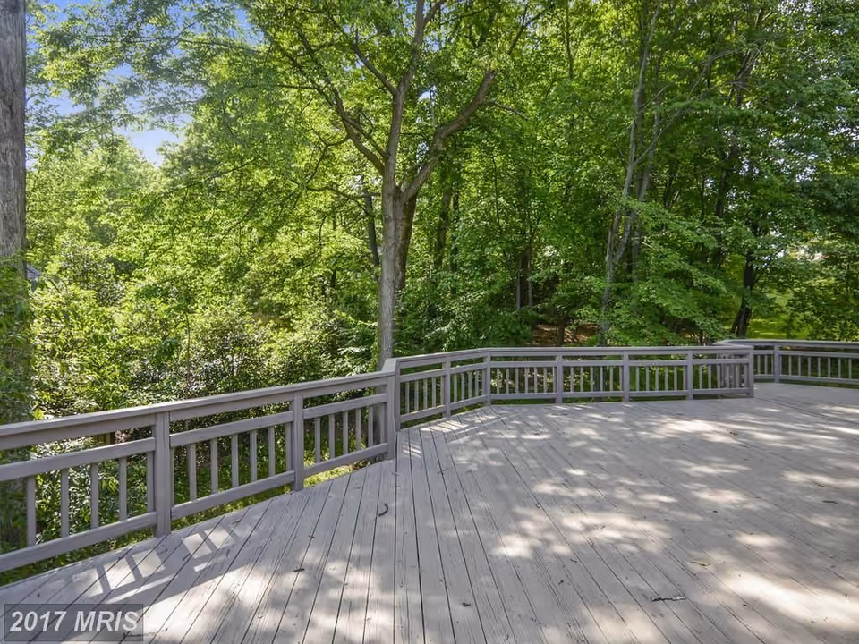 A spacious wooden deck with railings surrounded by lush green trees and foliage under a clear blue sky.