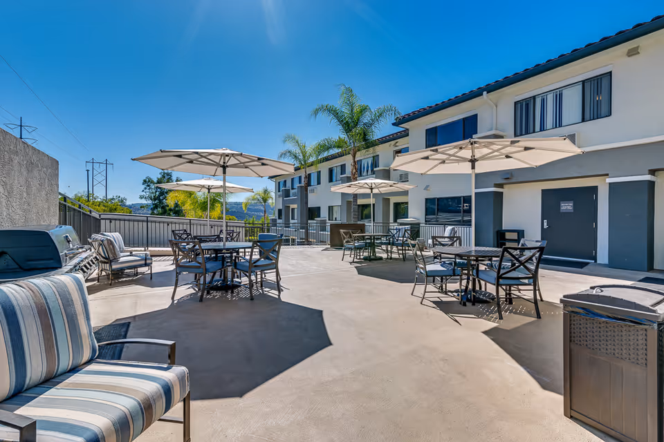 Outdoor patio with tables, umbrellas, and lounge chairs in front of a two-story senior living building under a clear blue sky.