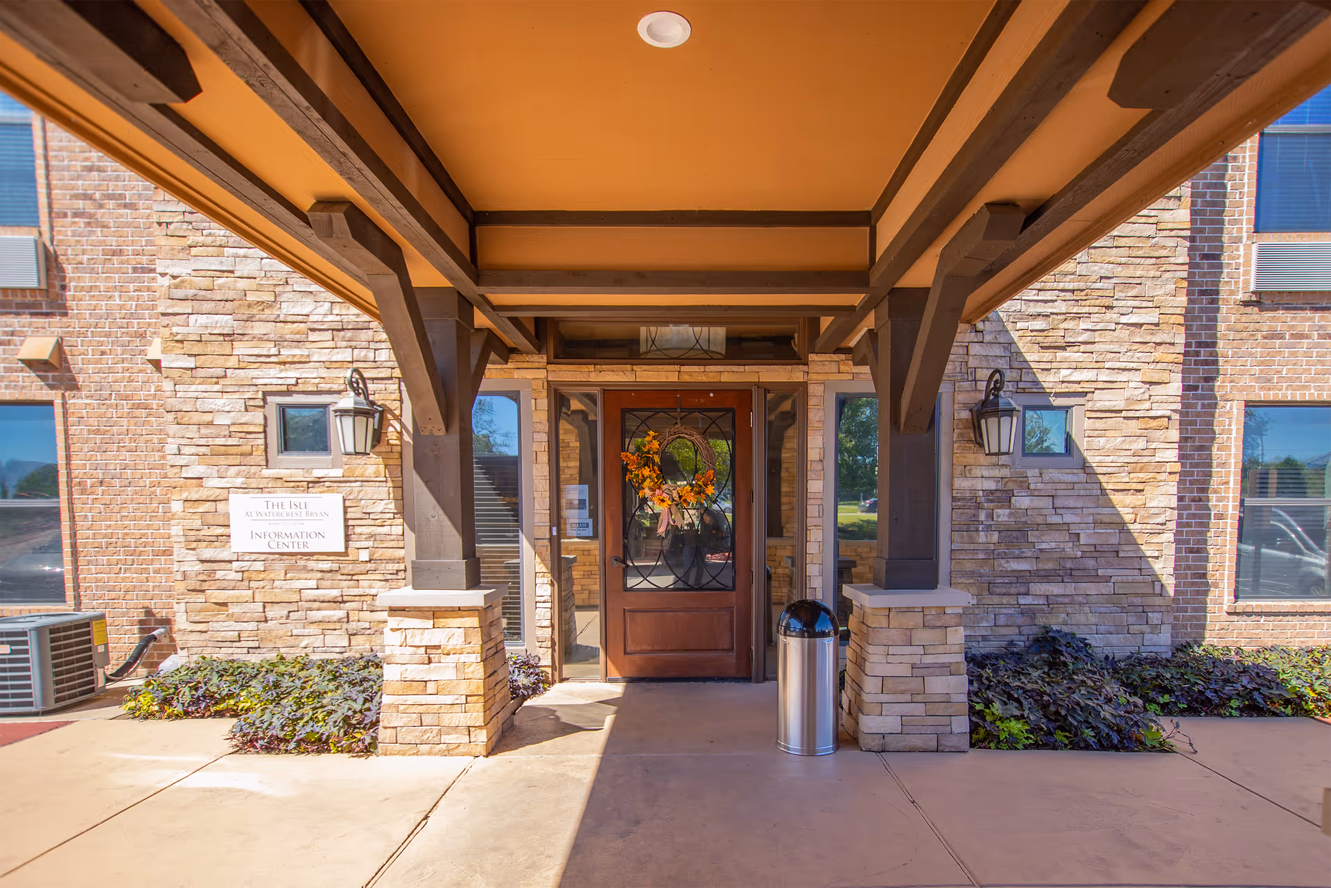 Entrance to The Isle at Watercrest Bryan Information Center with a wooden door decorated with a fall wreath, stone pillars, and a covered walkway.