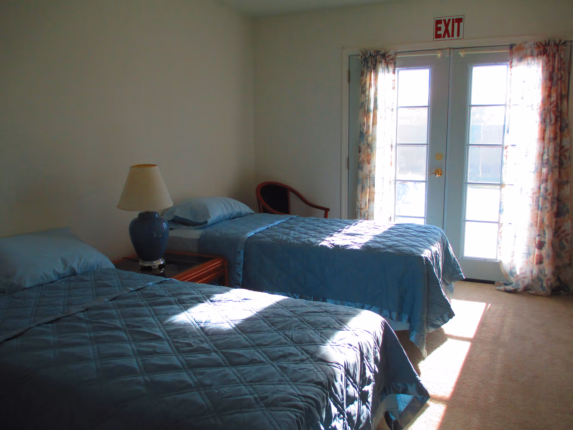Sunlit bedroom with two twin beds covered in blue quilts, a bedside table and lamp, and glass French doors topped by an EXIT sign.