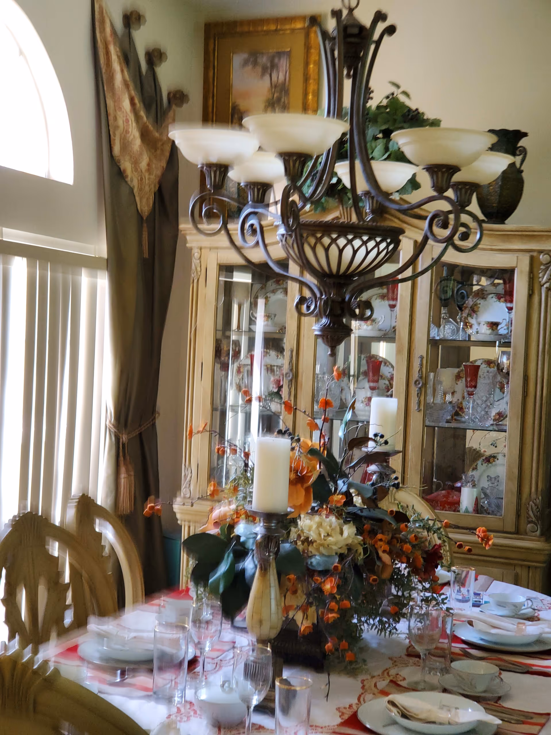 A dining room with a table set for a meal, featuring plates, glasses, and napkins. The table has a floral centerpiece with orange and white flowers and candles. Behind the table is a large wooden china cabinet displaying decorative plates and glassware. A chandelier with multiple lights hangs above the table, and there is a window with vertical blinds and curtains on the left side.