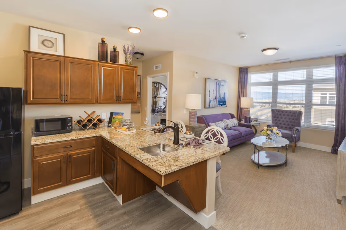 A bright and cozy living space featuring a kitchen area with wooden cabinets, granite countertops, a black refrigerator, and a microwave. The kitchen counter has a sink and is set with dishes and wine glasses. Adjacent to the kitchen is a living room with a purple sofa, a purple armchair, a round glass coffee table with a flower arrangement, and large windows with blinds and purple curtains letting in natural light.