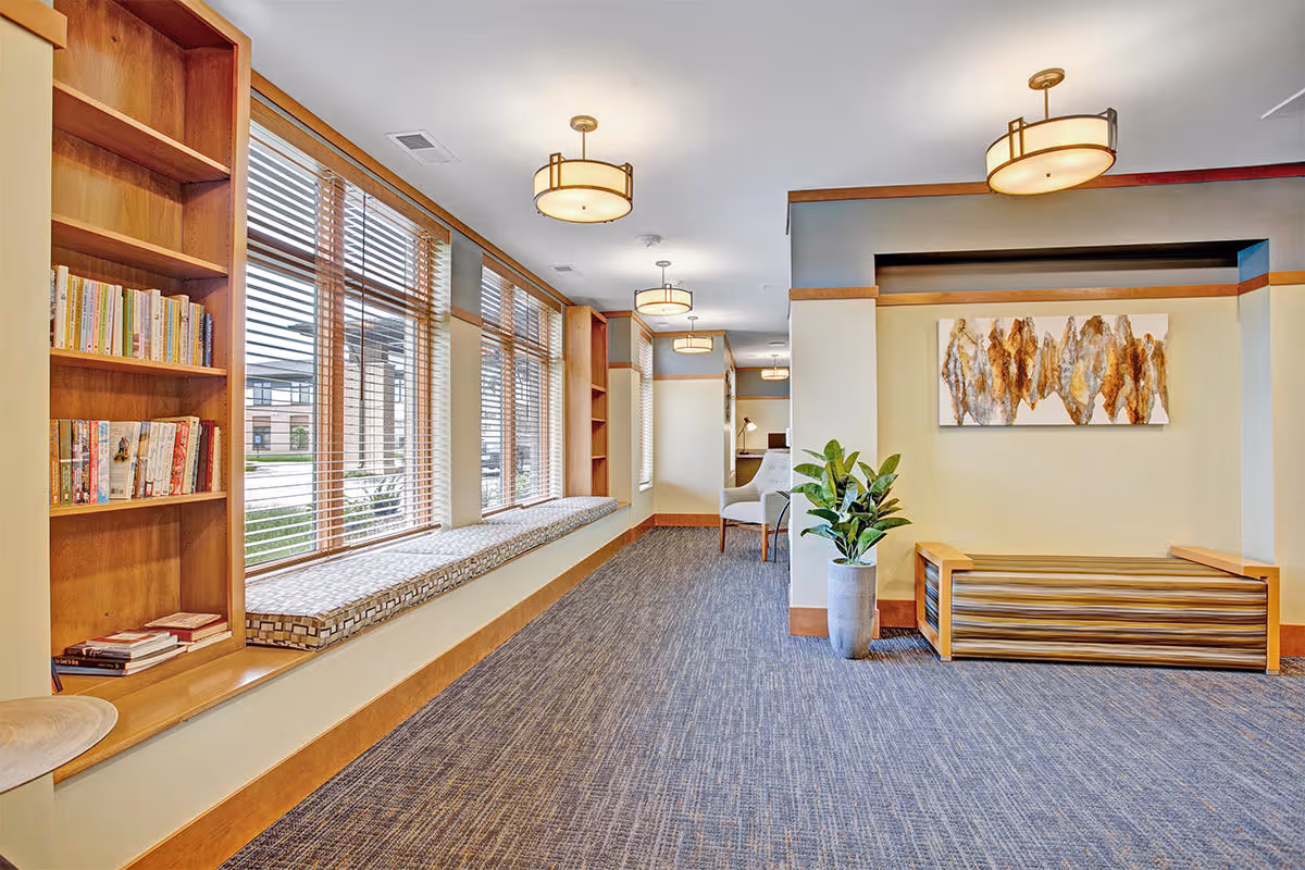 Bright common lounge with built-in bookshelves, window bench seating, pendant lights, a potted plant, and wall art.