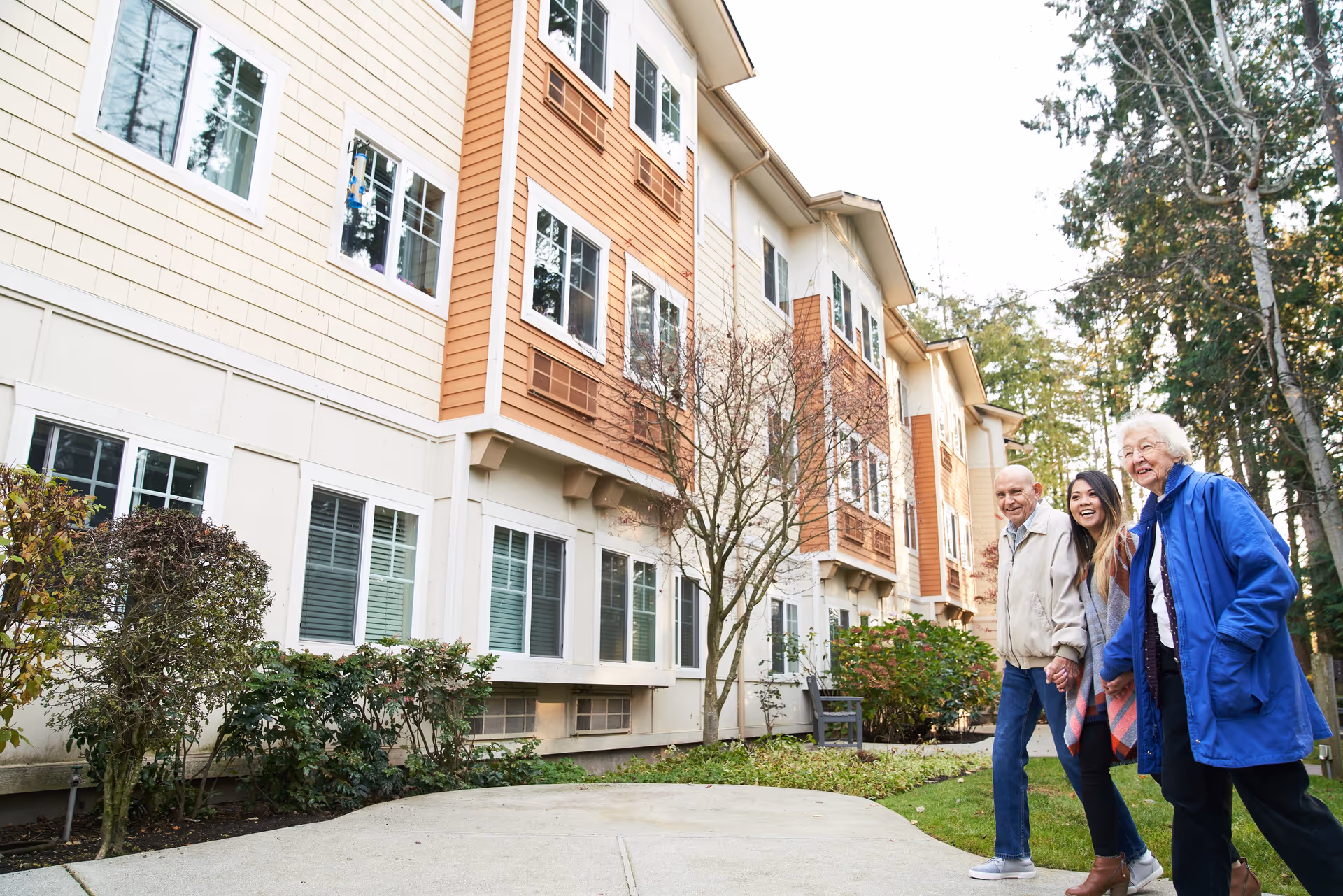 Three people, two elderly and one younger woman, walking together outside near a multi-story residential building with beige and brown siding, surrounded by trees and bushes.