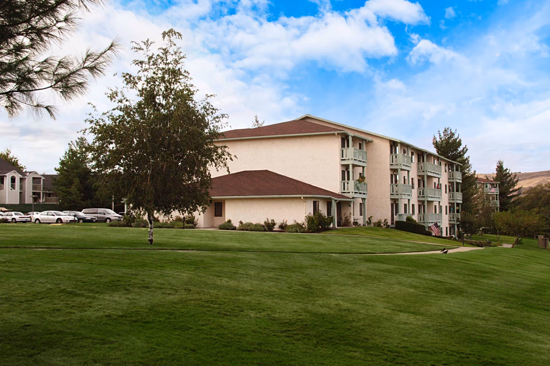 Exterior view of a multi-story beige senior living building with balconies, a wide green lawn, trees, and parked cars.