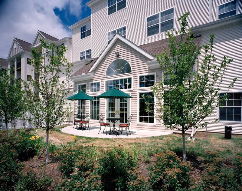 Outdoor patio with tables, chairs and green umbrellas in front of a multi-story white siding building.