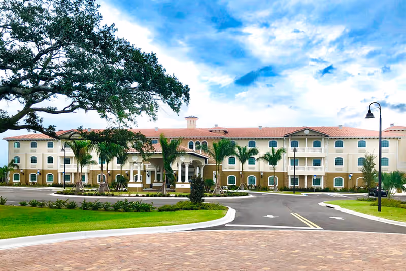 Front exterior view of a large, three-story senior living facility building with a red-tiled roof, multiple windows, balconies, palm trees, and a circular driveway under a partly cloudy blue sky.