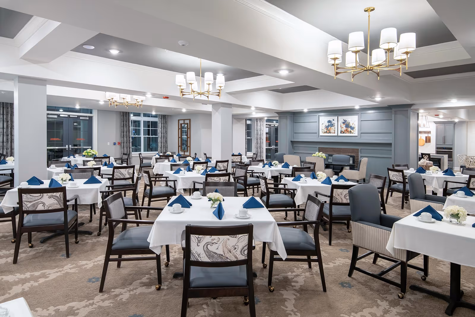A spacious senior living dining room with multiple tables covered in white tablecloths, each set with blue folded napkins, white cups, and small floral centerpieces. The room features comfortable chairs with patterned backs and cushioned seats, elegant chandeliers hanging from the ceiling, large windows with curtains, and a decorative fireplace with framed artwork above it.
