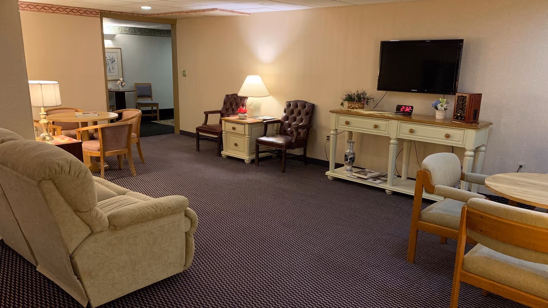A cozy living room area with a beige recliner sofa on the left, two brown leather armchairs with a small table and lamp between them against the back wall, and a white console table with a TV mounted above it. There are two round wooden tables with chairs on the right side of the room. The carpet is dark patterned, and the walls are light-colored. The room is softly lit with lamps and ceiling lights.