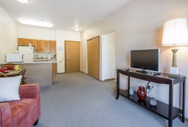 A living area in a senior living facility featuring a red armchair with a white pillow, a dark wooden TV stand with a flat-screen television, decorative vases, and a lamp. In the background, there is a small kitchen area with wooden cabinets, a refrigerator, microwave, and a counter with two chairs. The room has beige walls and carpeted flooring, with two wooden doors visible.