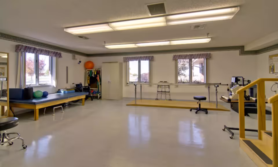 A spacious physical therapy or rehabilitation room with exercise equipment including parallel bars, therapy tables, exercise balls, and stools. The room has large windows with purple valances allowing natural light to enter, and fluorescent ceiling lights providing additional illumination.
