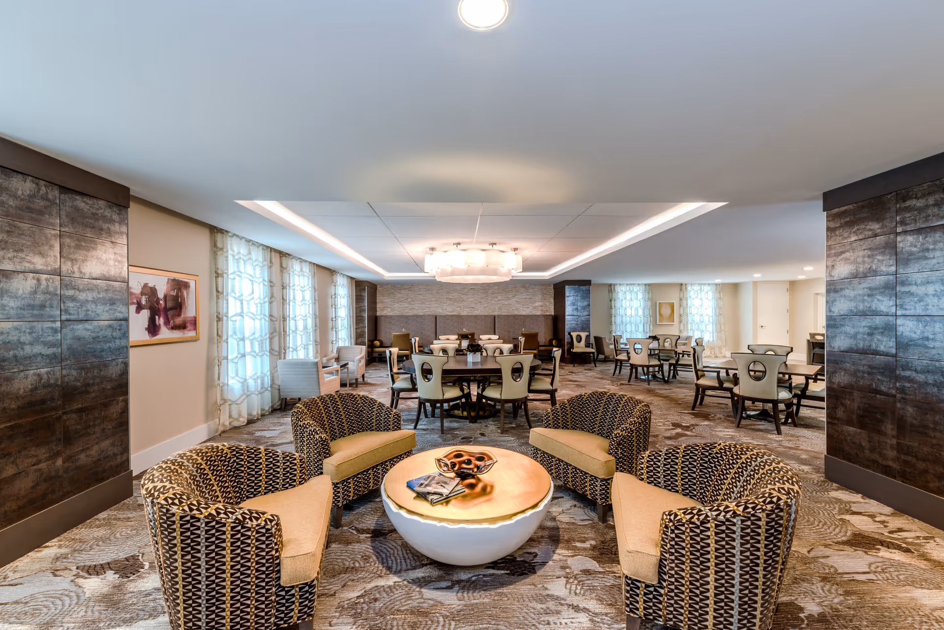 A spacious and well-lit common area with patterned carpet flooring, featuring four cushioned armchairs arranged around a round coffee table with decorative items on top. In the background, there are multiple dining tables with chairs, large windows with sheer curtains, and modern wall decor. The ceiling has recessed lighting and a central chandelier.