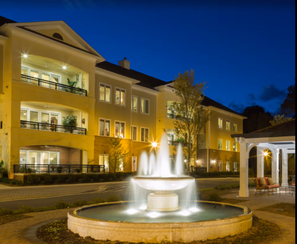 Nighttime exterior of a three-story senior living building with illuminated balconies and a central fountain in the foreground.