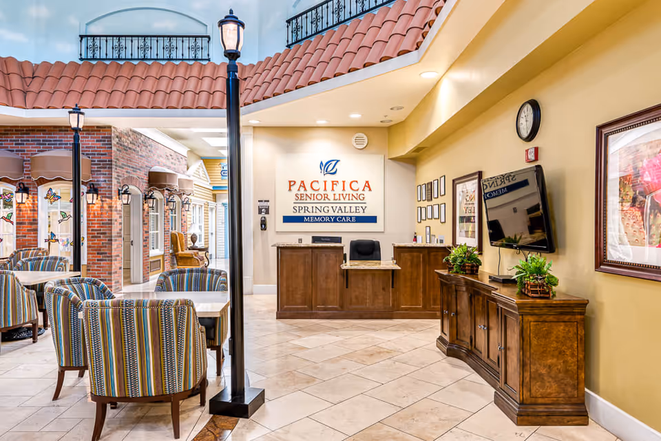 Interior view of the reception area at Pacifica Senior Living Spring Valley Memory Care, featuring a wooden reception desk, a wall sign with the facility name, a TV mounted on the wall, framed artwork, and a seating area with striped upholstered chairs and tables. The space has a tiled floor, warm lighting, and decorative streetlamp-style lights.