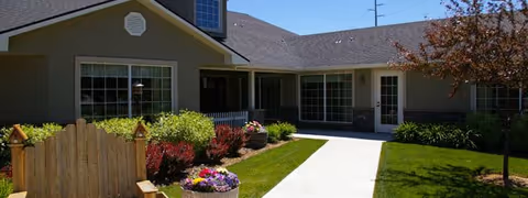 Front exterior of a single-story assisted living cottage with a paved walkway, green lawn, and flower beds.