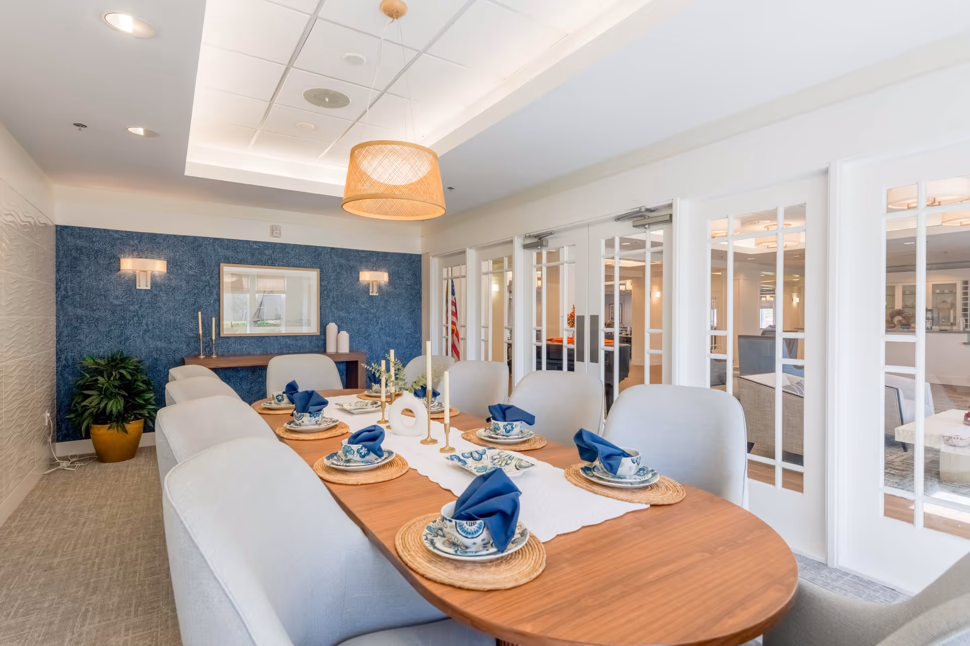 A modern dining room with a wooden table set for six, featuring blue napkins folded on patterned plates placed on woven placemats. The room has light gray upholstered chairs, a blue accent wall with two wall sconces, a framed picture, and a potted plant. There are white double doors with glass panels leading to another room with seating visible through the doors. A woven pendant light hangs from the ceiling.
