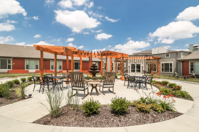 Outdoor courtyard area at Carnegie Village Senior Living Community featuring a circular paved patio with wooden pergolas, metal tables and chairs, wooden lounge chairs, a central water fountain, and surrounding landscaped garden beds under a partly cloudy blue sky.