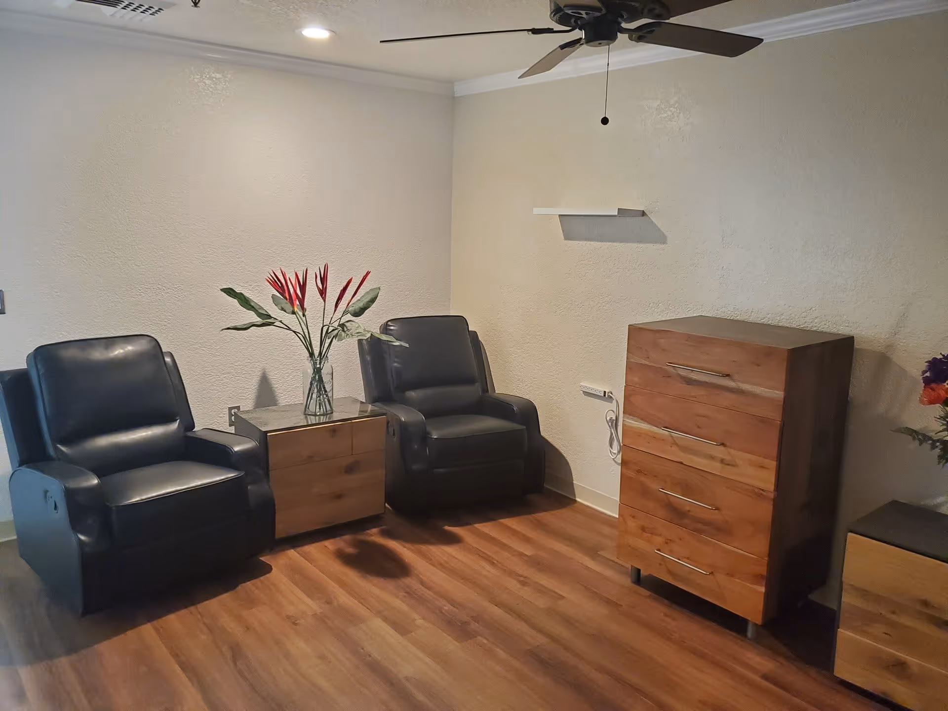 Small sitting area with two black leather recliners flanking a wooden side table with a vase of red flowers, a wooden chest of drawers, and a ceiling fan in a room with hardwood floors.