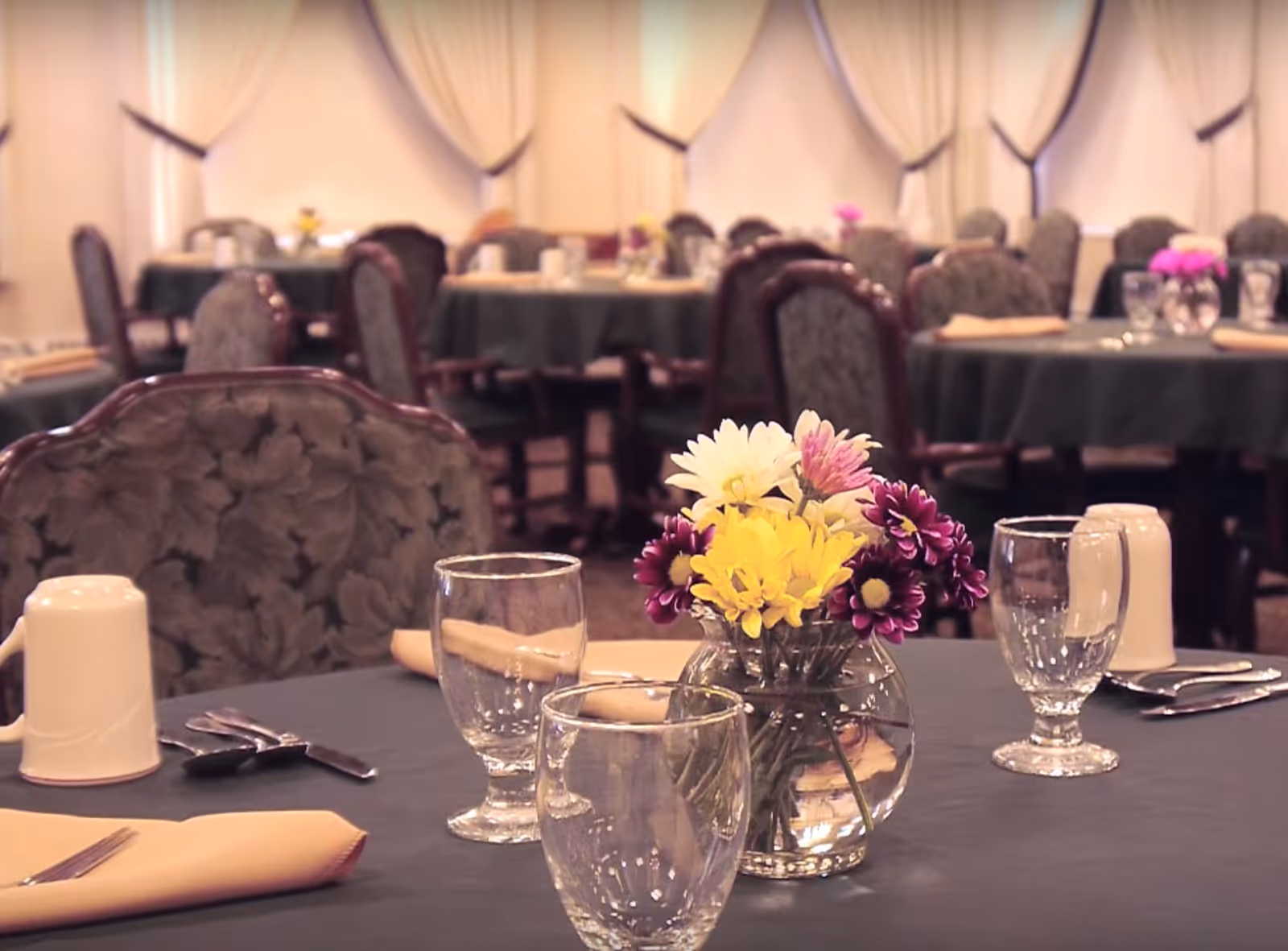 Dining room table set with glassware, napkins, silverware and a small vase of yellow and purple flowers, with more tables and chairs in the background.