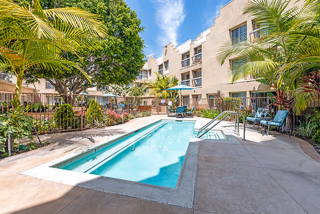 Outdoor courtyard with a rectangular swimming pool, lounge chairs and umbrellas, surrounded by a multi-story apartment building and tropical landscaping.