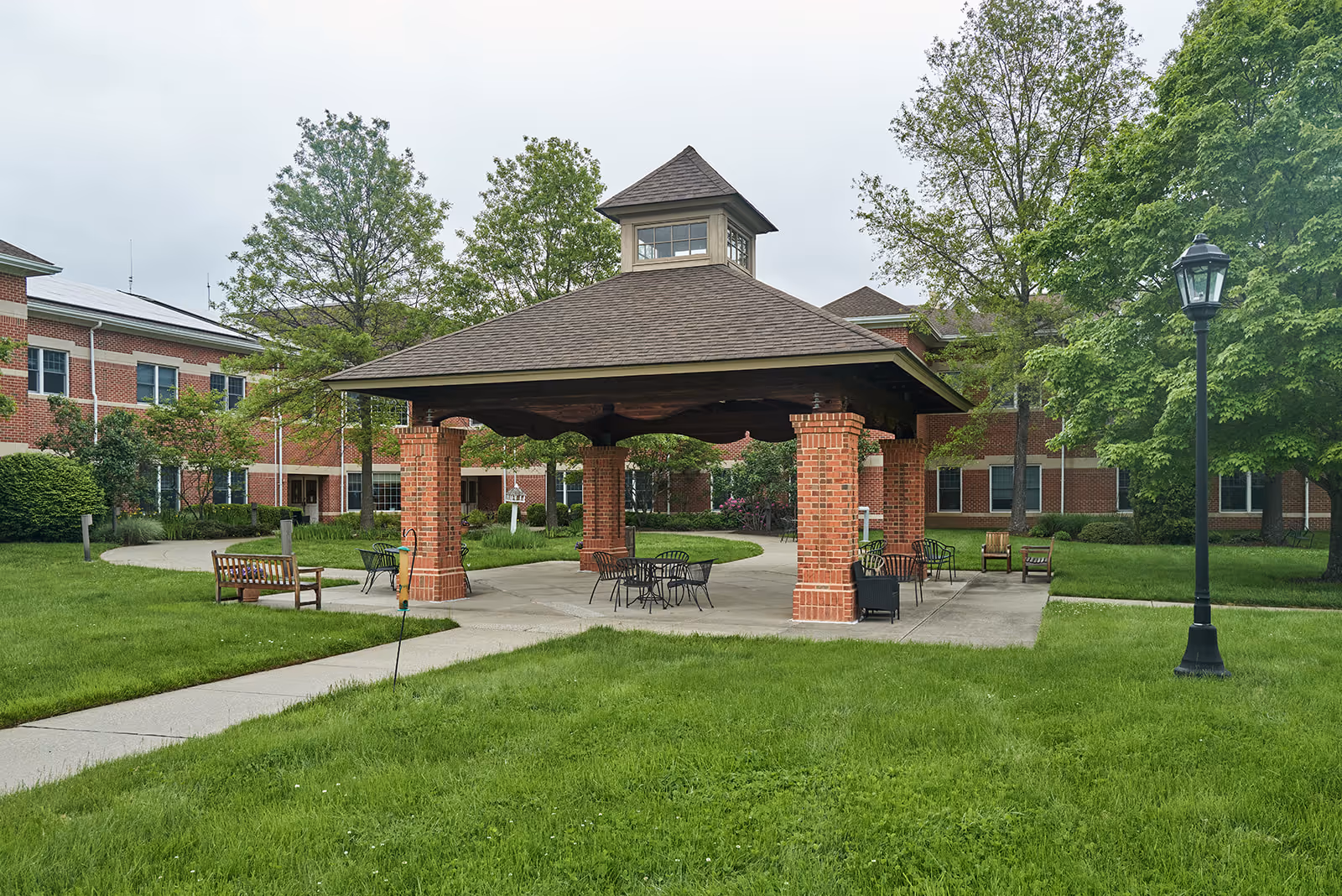 Covered brick pavilion with tables and benches in a grassy courtyard in front of a multi-story brick senior living building.